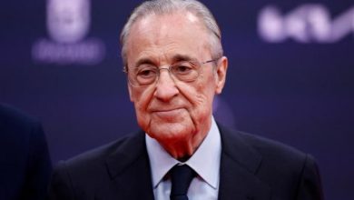 Older man in a dark suit and tie, wearing glasses, looking slightly downward with a neutral expression at a formal event.