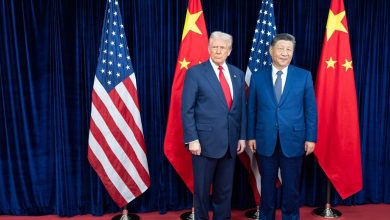 Two men in dark suits stand side by side on a red carpet with U.S. and Chinese flags behind them.