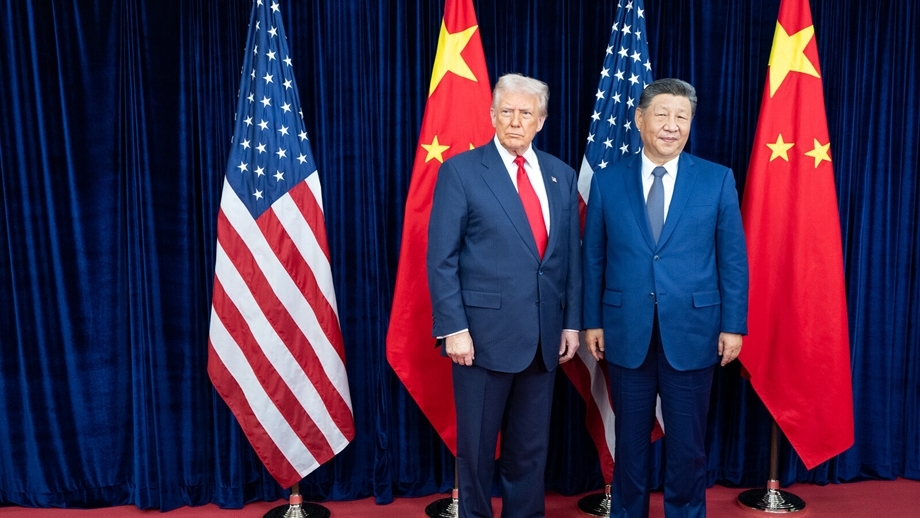 Two men in dark suits stand side by side on a red carpet with U.S. and Chinese flags behind them.