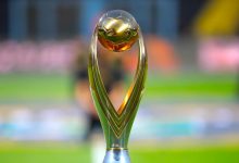 vis_0522202622133390 | MCG24 | موقع إلكتروني مغربي شامل متجدد على مدار الساعة Gold sports trophy with a round golden ball atop a curved green base on a blurred stadium field in the background, symbolizing victory.