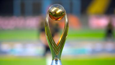 Gold sports trophy with a round golden ball atop a curved green base on a blurred stadium field in the background, symbolizing victory.