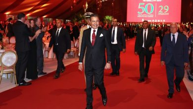vis_0522202623003833 | MCG24 | موقع إلكتروني مغربي شامل متجدد على مدار الساعة Smiling man in a dark suit and maroon tie walks the red carpet at a formal gala, with guests and a large red screen behind.