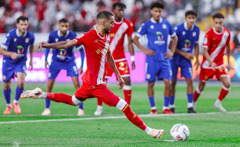 vis_0523202621270270 | MCG24 | موقع إلكتروني مغربي شامل متجدد على مدار الساعة Red-clad soccer player explodes into a kick toward the ball while defenders in blue watch from behind on a sunny pitch.