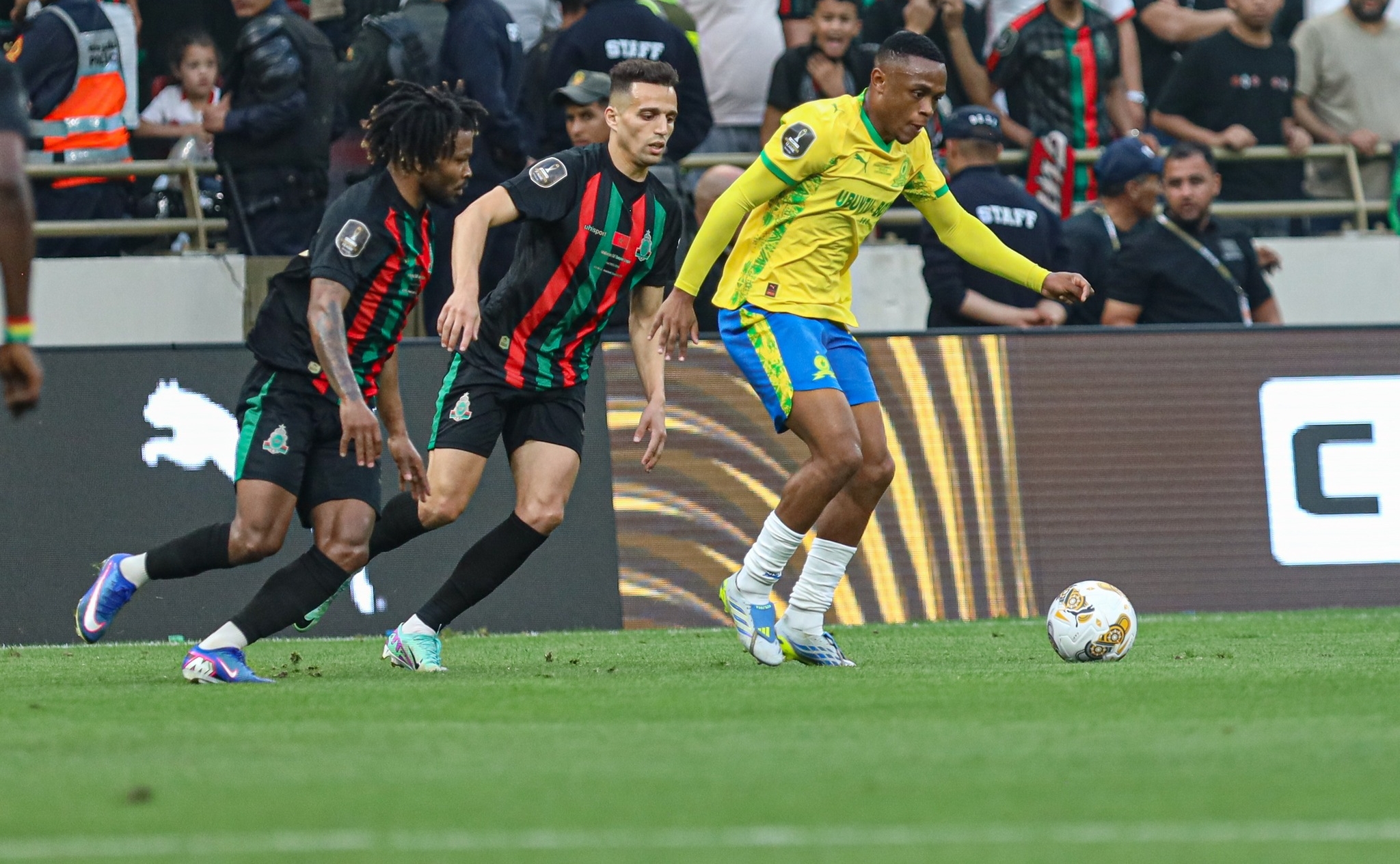 Soccer players chase the ball on a green pitch with spectators in the stands watching from behind the goal area.