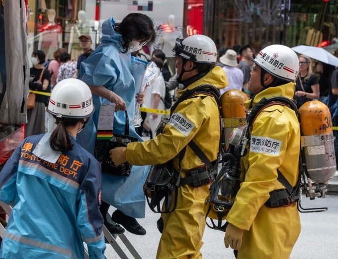 Firefighters in yellow suits and helmets assist a person in a blue protective gown on a crowded street.
