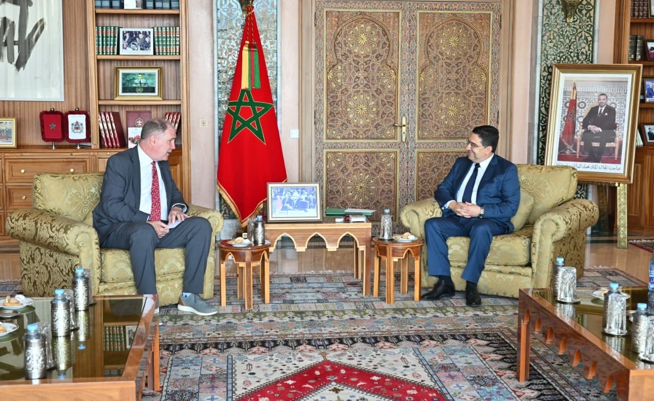 vis_0525202617422357 | MCG24 | موقع إلكتروني مغربي شامل متجدد على مدار الساعة Two men in suits sit on ornate sofas across from each other in a formal office, Moroccan flag between them.