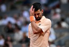 Male tennis player in peach shirt touching his forehead with his right hand on the court, orange wristband on his wrist, blurred crowd in background.