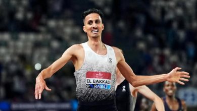 vis_0531202621223713 | MCG24 | موقع إلكتروني مغربي شامل متجدد على مدار الساعة Male track athlete in a white gradient singlet with a red El Bakkali bib, arms outstretched in celebration on a stadium track.
