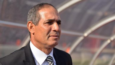 2456688 | MCG24 | موقع إلكتروني مغربي شامل متجدد على مدار الساعة Middle-aged man in a black suit and striped tie stands outdoors, looking to the right near a curved shelter or dugout.