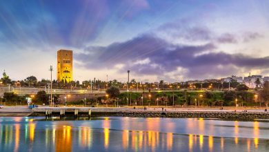 Sunset over a riverside with a tall stone tower, palm trees, and a promenade reflecting warm lights on the water.