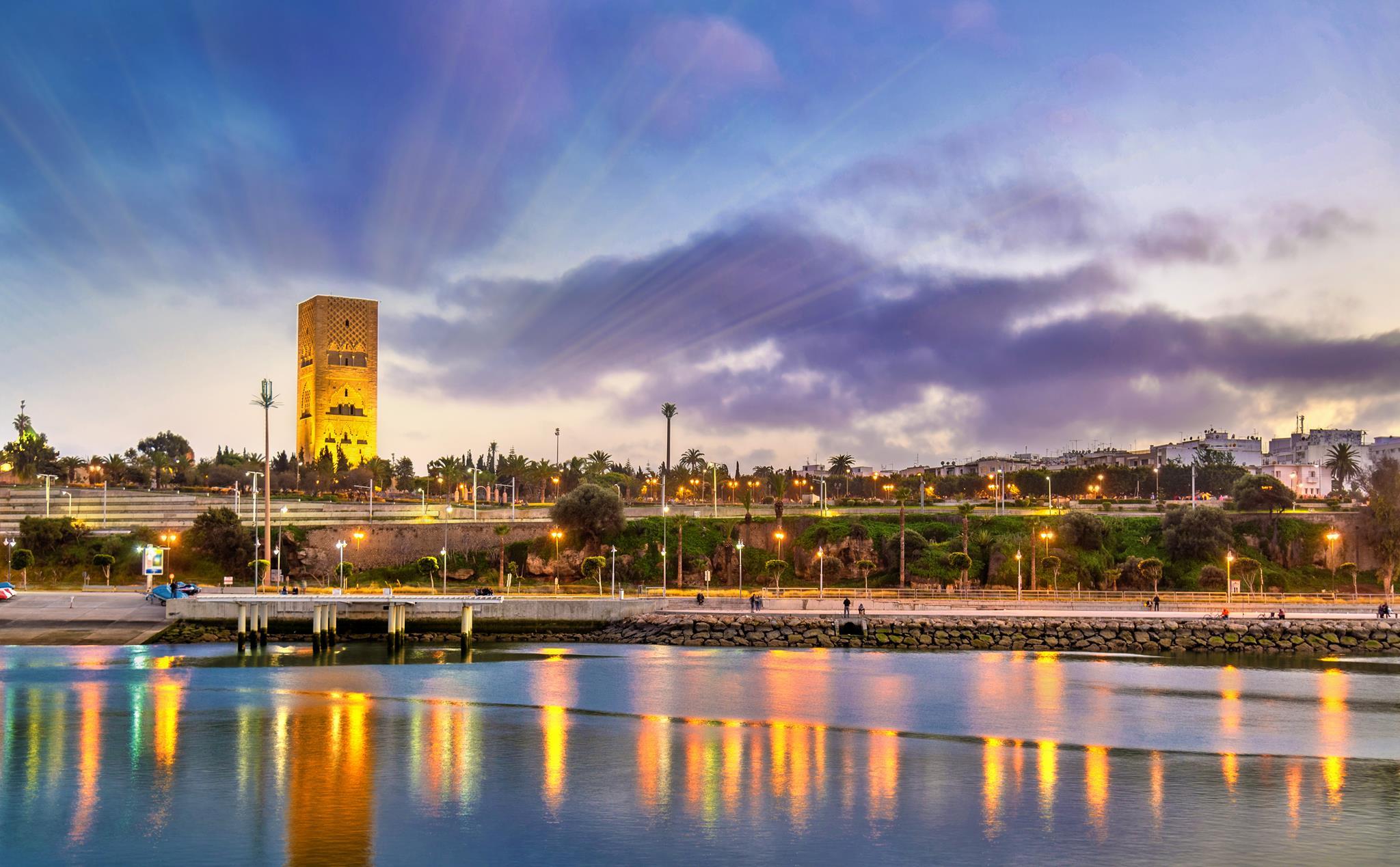 Sunset over a riverside with a tall stone tower, palm trees, and a promenade reflecting warm lights on the water.