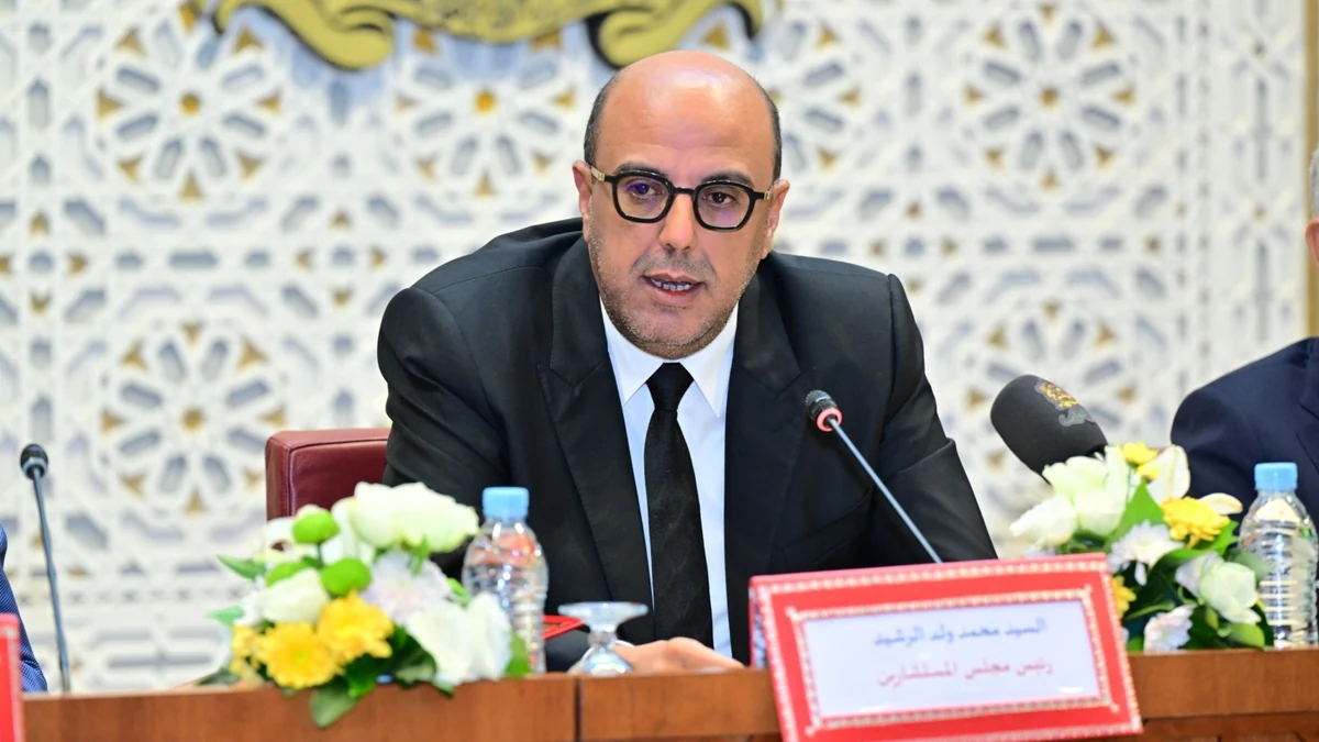 Man in a black suit and glasses speaks at a conference, seated behind a wooden table with flowers and microphones in front of him.