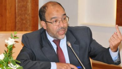 أحمد-صدقي | MCG24 | موقع إلكتروني مغربي شامل متجدد على مدار الساعة Man in a dark suit and red tie speaking into a microphone at a panel, gesturing with his right hand while seated at a table with flowers nearby.