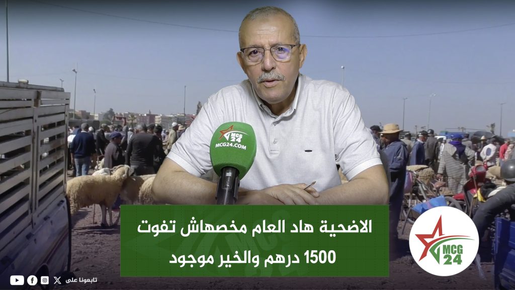 News presenter speaking into a green MC24 microphone, seated at a desk with a crowded market background and Arabic headline overlay.