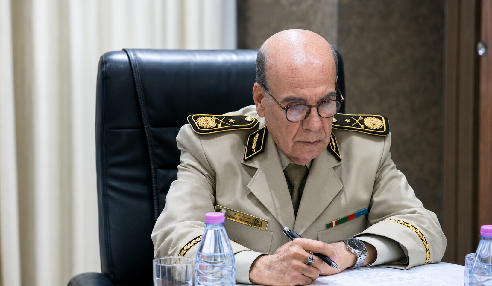 Senior military officer in a beige dress uniform, writing at a desk with a pen, wearing glasses and a watch.