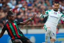 Two soccer players clash for the ball during a match: one in a black kit with red and green stripes challenges a white-and-green player.