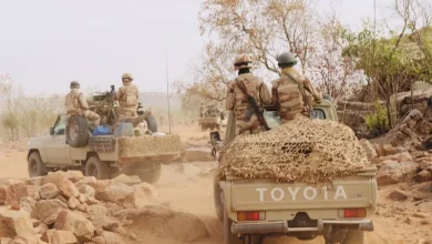 Soldiers in desert camouflage patrol in open-top Toyota pickups with netting, driving along a dusty dirt road.