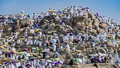 Large crowd of people in white clothing gathered on a rocky hill, many holding colorful umbrellas against a blue sky.