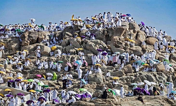 Large crowd of people in white clothing gathered on a rocky hill, many holding colorful umbrellas against a blue sky.