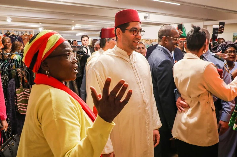 Group of people at an indoor cultural event; foreground left shows a woman in yellow with a red headwrap gesturing as a man in white traditional attire and a red fez watches nearby, with others in the background.