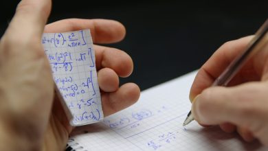 Hands holding a torn notebook page filled with blue handwritten equations, while writing on graph paper with a pen.