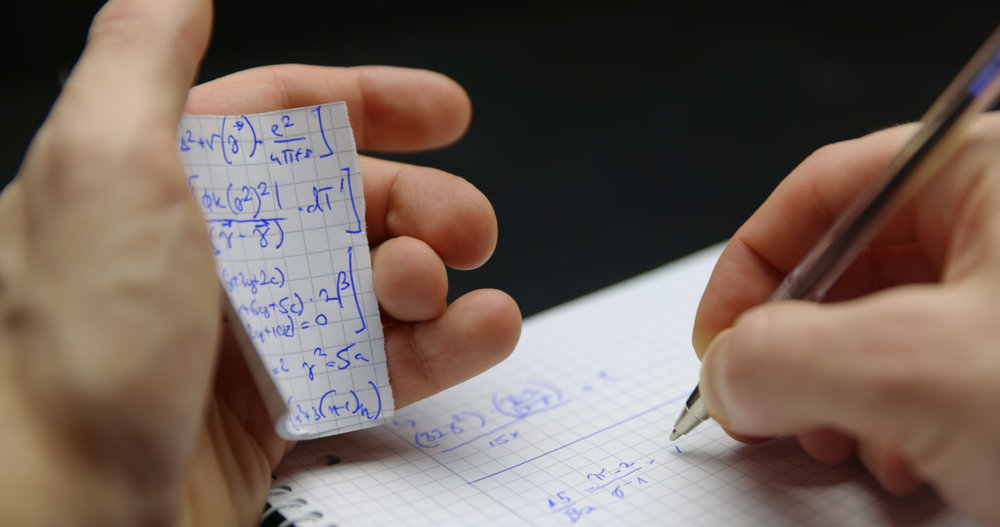 Hands holding a torn notebook page filled with blue handwritten equations, while writing on graph paper with a pen.