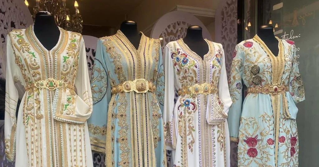 القفطان-المغربي | MCG24 | موقع إلكتروني مغربي شامل متجدد على مدار الساعة Group of four embellished, pastel-colored dresses on mannequins with gold embroidery and jeweled belts in a shop window display.