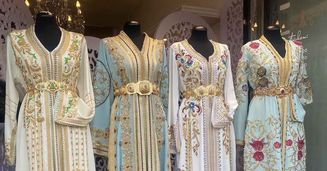Group of four embellished, pastel-colored dresses on mannequins with gold embroidery and jeweled belts in a shop window display.