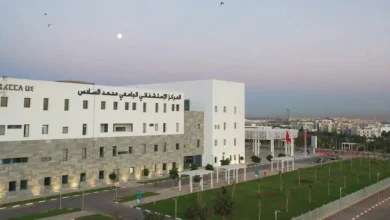 White and gray multi-story medical center with Arabic signage, stone-clad lower facade, and a manicured lawn along a road at dusk.