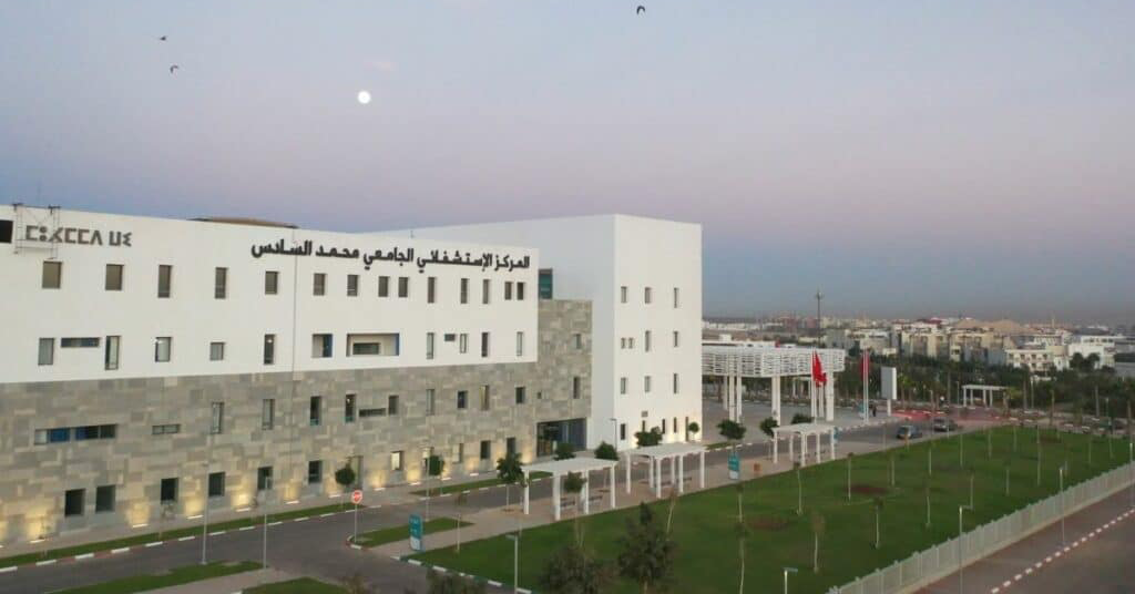 White and gray multi-story medical center with Arabic signage, stone-clad lower facade, and a manicured lawn along a road at dusk.