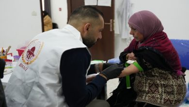 Healthcare worker wearing a white vest administers a vaccination to a seated woman wearing a pink hijab in a clinic setting.