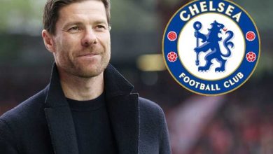 Man in a dark coat outdoors with the Chelsea Football Club logo beside him.