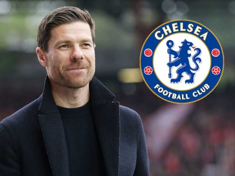 Man in a dark coat outdoors with the Chelsea Football Club logo beside him.