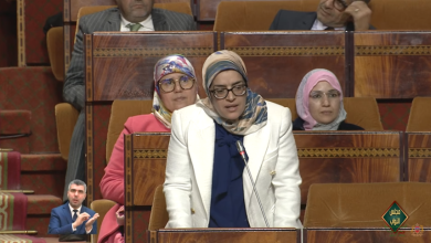 Woman in a white blazer speaks at a microphone in a parliamentary chamber, with three women in hijabs seated behind in the burgundy seating area, a sign-language interpreter inset at lower left.