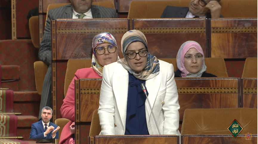 ثورية-عفيف | MCG24 | موقع إلكتروني مغربي شامل متجدد على مدار الساعة Woman in a white blazer speaks at a microphone in a parliamentary chamber, with three women in hijabs seated behind in the burgundy seating area, a sign-language interpreter inset at lower left.