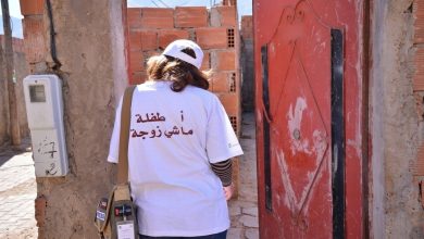 Person wearing a white T‑shirt with Arabic text on the back, carrying a tan shoulder bag and press badge, walking through a brick-walled entry beside a red door.
