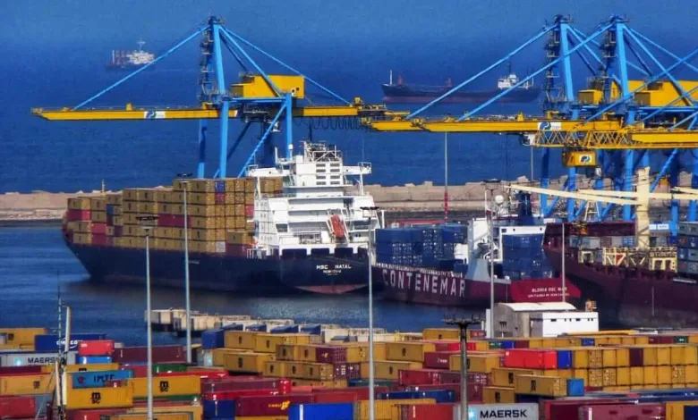 Container port with blue gantry cranes loading a cargo ship; colorful containers stacked on the dock and ships at sea in the background.