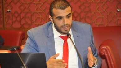 Man in a blue suit and red tie speaks into a microphone during a panel discussion, gesturing with his hands beside a laptop.