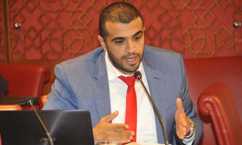 Man in a blue suit and red tie speaks into a microphone during a panel discussion, gesturing with his hands beside a laptop.