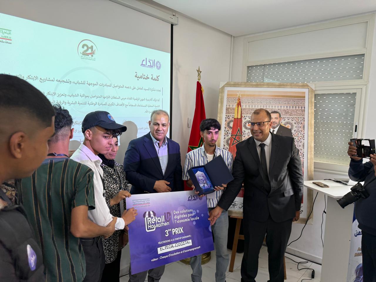 Group of people at an award ceremony; a young man receives a certificate and a purple prize banner while officials smile, with a Moroccan flag in the background.