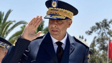 Male military officer in dress uniform saluting, wearing a dark blue peaked cap with gold braid, outdoors with palm trees in the background.