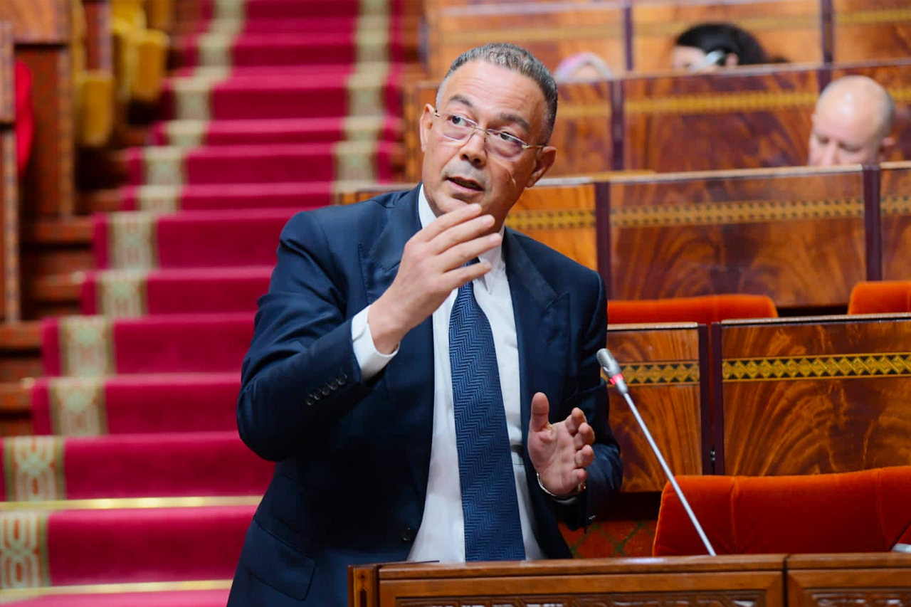 A man in a dark suit and blue tie speaks at a podium in a wooden parliamentary chamber with red-carpet steps behind him.