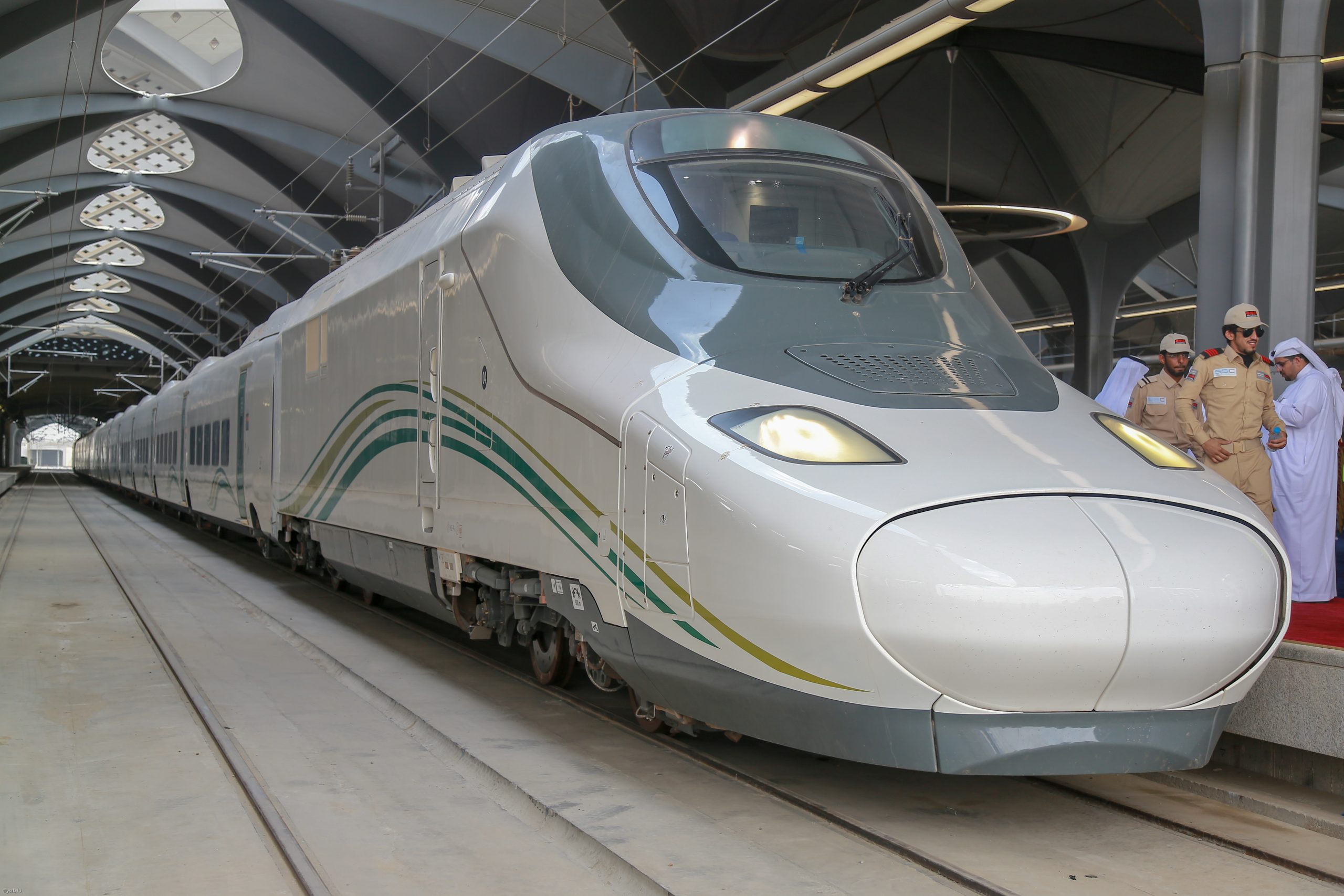 High-speed silver train at a station with engineers in beige uniforms and traditional white garments nearby in the background? (informative)