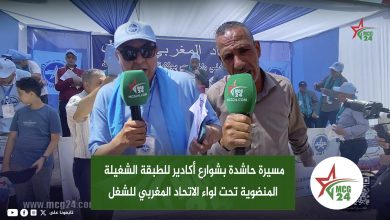 Two men holding green MCG24 microphones conduct an on-site interview at a tented event, with a blue backdrop banner and onlookers in the background.