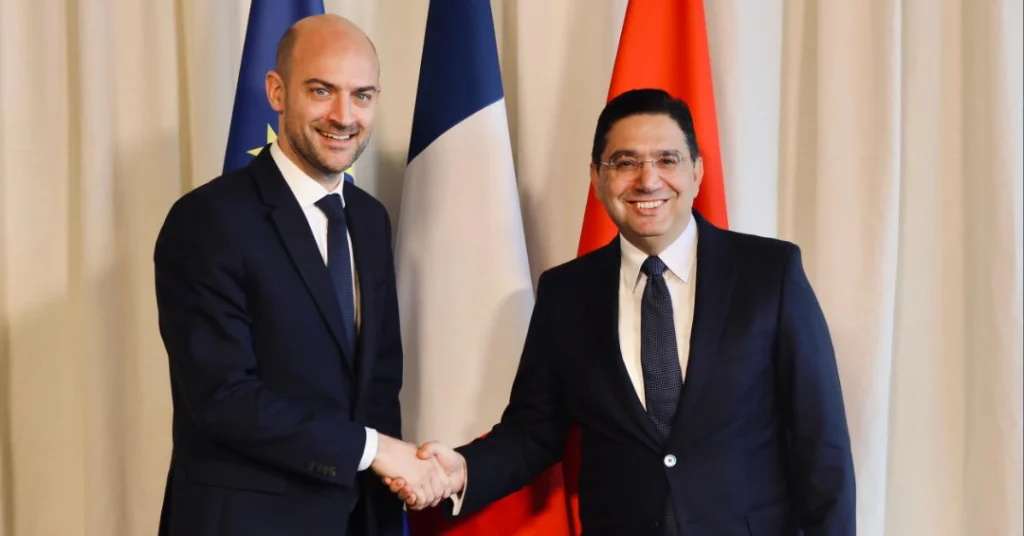 Two men in dark suits shake hands in front of three vertical flags.