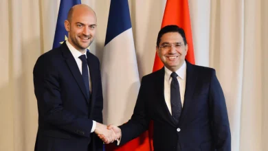 Two men in dark suits shake hands in front of three vertical flags.