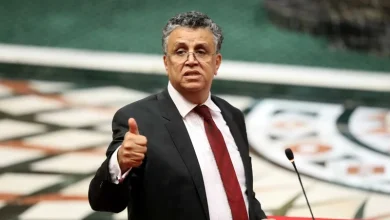Man in a dark suit and red tie gives a thumbs-up at a podium with a microphone in a formal setting.