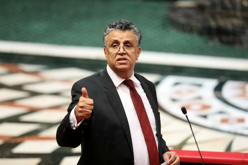 Man in a dark suit and red tie gives a thumbs-up at a podium with a microphone in a formal setting.