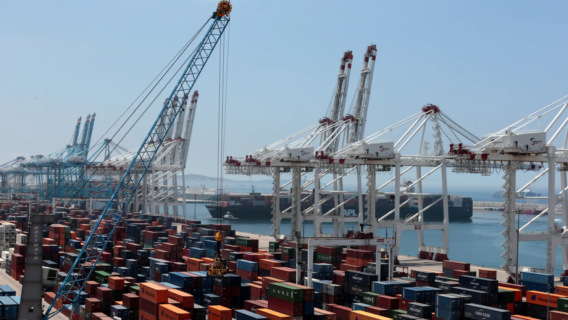 00032-1712425416 | MCG24 | موقع إلكتروني مغربي شامل متجدد على مدار الساعة Port scene with tall gantry cranes over stacked shipping containers and a cargo ship in the background by the water in a busy harbor.