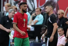 Two male players converse on a sports court; one in a red jersey with number 3 and green shorts, the other in black.